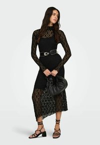 Černé krajkové šaty s dlouhými rukávy, vysokým límcem, po střed lýtek a průhlednými detaily. Černý pásek s hroty a černá volná kabelka; sandály na tenkých páscích.
