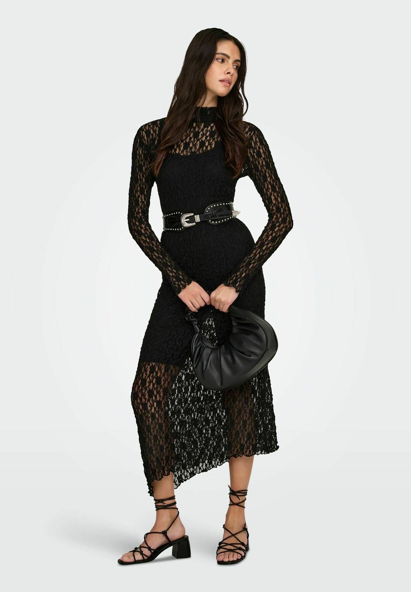 Černé krajkové šaty s dlouhými rukávy, vysokým límcem, po střed lýtek a průhlednými detaily. Černý pásek s hroty a černá volná kabelka; sandály na tenkých páscích.