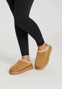 Chaussons en suède beige avec une bordure en fausse fourrure crème, présentant une semelle en caoutchouc texturé et un design à bout rond. Portés avec des leggings noirs.