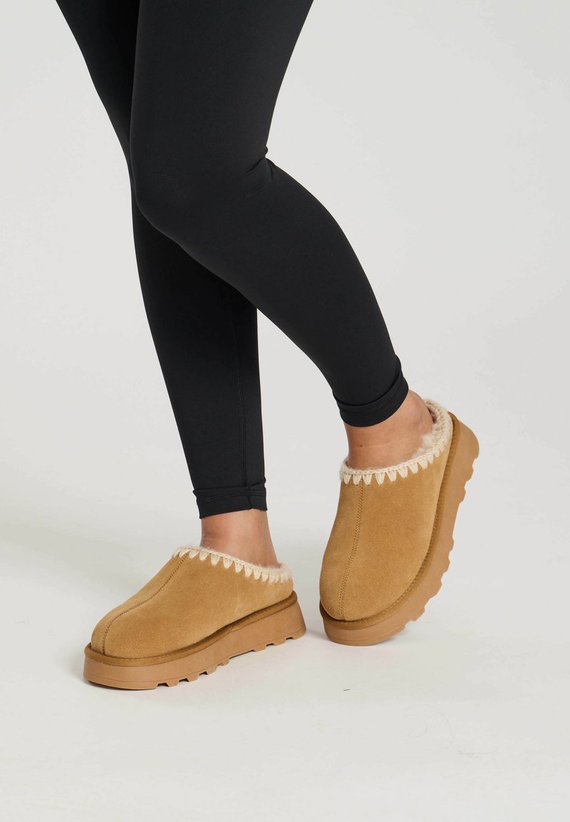 Chaussons en suède beige avec une bordure en fausse fourrure crème, présentant une semelle en caoutchouc texturé et un design à bout rond. Portés avec des leggings noirs.