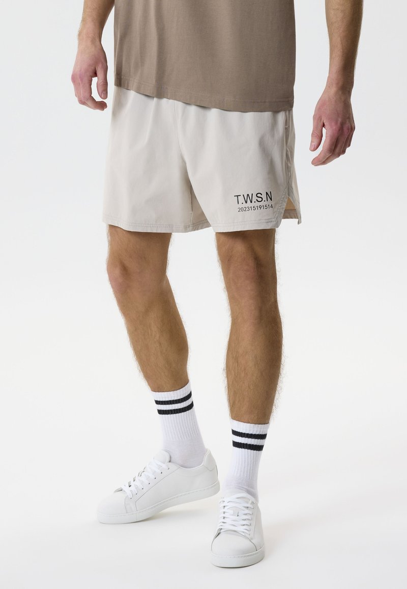 Mann trägt beige Shorts mit dem Schriftzug "T.W.S.N", weiße Sneaker und weiße Socken mit zwei schwarzen Streifen, steht vor einem einfarbigen Hintergrund.