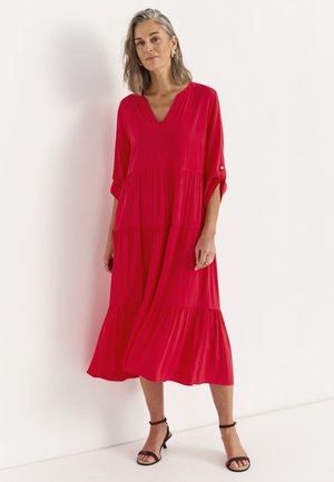 Kvinde med skulderlangt gråt hår iført en løs, lys rød midi-kjole med opkørte ærmer og sorte ankelrem-sandaler.