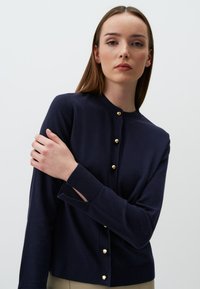 Navyfarbener Cardigan aus weichem Material mit Rundhalsausschnitt, goldenen Knöpfen und Ausschnitten an den Manschetten. Schlichtes, klassisches Design.