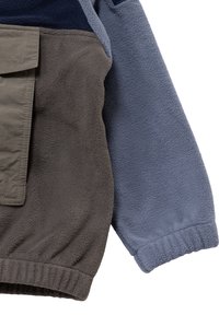 Kurtka polarowa w szare i granatowe sekcje, z dużą brązową kieszenią z przodu, ściągaczami na mankietach i miękką teksturą.