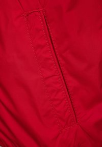 Roter Jackenstoff mit einer vertikalen Reißverschlusstasche auf der linken Seite. Die Textur ist glatt und die Nähte sind entlang der Nähte sichtbar.