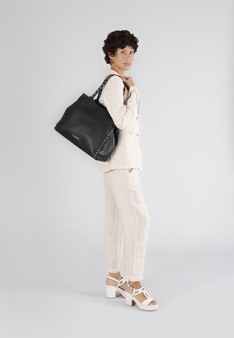 Borsa tote in pelle nera con dettagli in borchie, dotata di due manici e un design spazioso. Il modello indossa un completo a righe chiare e scarpe bianche.