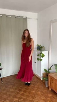 Robe longue, sans bretelles, de couleur bordeaux avec une jupe fluide. Le tissu semble léger avec des plis verticaux et un éclat subtil. Talons noirs.