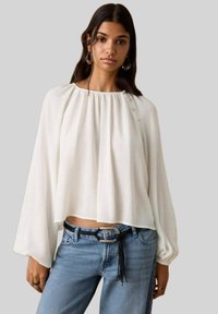 Weiße, leichte Bluse mit gerafftem Ausschnitt, langen Ärmeln und entspanntem Schnitt, kombiniert mit hellblauen, hoch taillierten Jeans.