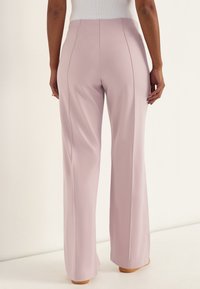 Frau trägt hoch taillierte, weit geschnittene hellrosa Hosen, steht mit entspannten Armen und ist von hinten vor neutralem Hintergrund zu sehen.