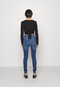 Svart långärmad topp med knytband bak, matchad med åtsittande mörkblå jeans och kraftiga svarta klackar mot en vit bakgrund.