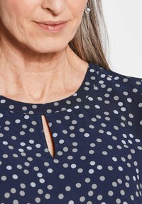 Marineblauwe shirt met een ronde halslijn en een sleutelgaatje, versierd met grijs-witte stippen in verschillende maten.
