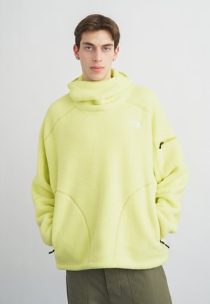 Mladić u žutom flis puloveru s visokim ovratnikom i rukama u džepovima, stoji ispred jednobojne svijetle pozadine.