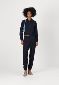 Marine gestreept tweedelig outfit van zachte stof, met een colbert met ritssluiting en tapered broek, gecombineerd met blauwe blokhak sandalen.