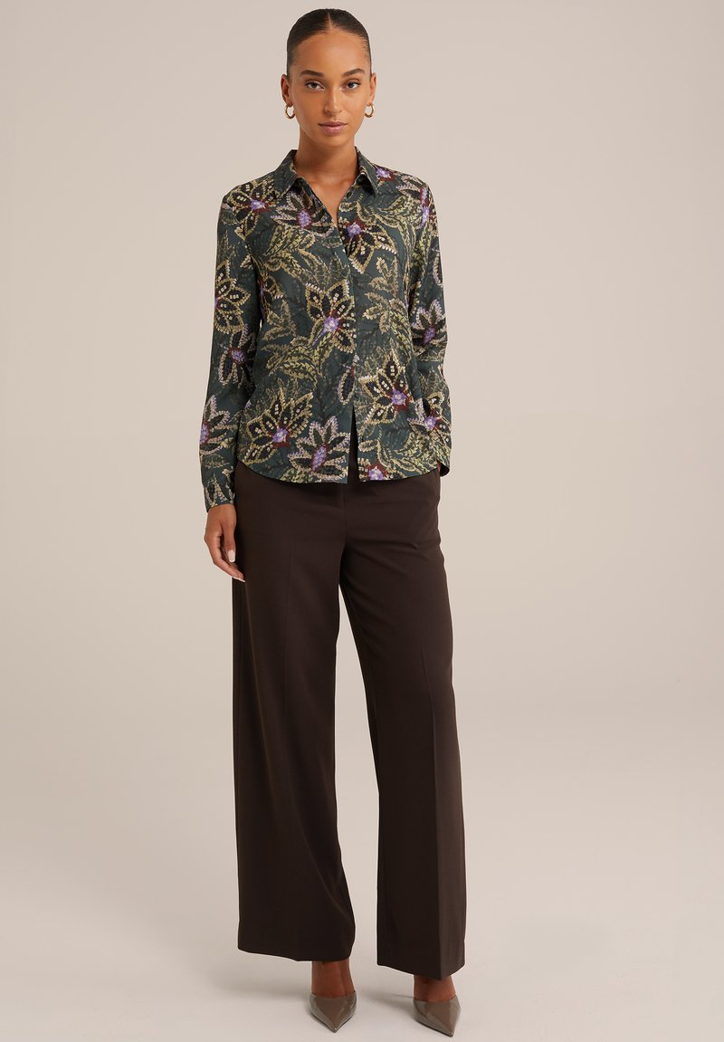 Geknoopte overhemd met kraag in een donkergroene stof, voorzien van een bloemenpatroon in paars en goud, gecombineerd met losse bruine broek en hakken.