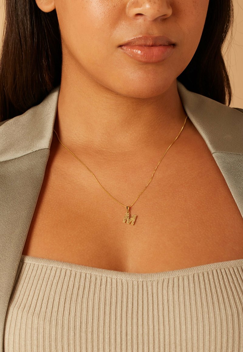 Gouden ketting met een textuur "M" hanger, gedragen op een geribd beige top. De ketting is dun en delicaat, wat het ontwerp van de hanger goed aanvult.