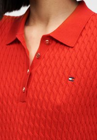 Κόκκινο πλεκτό πόλο πουκάμισο με υφασμένο σχέδιο, κουμπότρυπα και μικρό κεντημένο λογότυπο στο αριστερό στήθος.