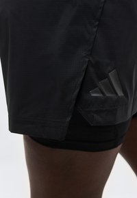 Μαύρο αθλητικό σορτς με επικαλυπτόμενο πλευρικό πάνελ και διακριτικό λογότυπο Adidas στο κάτω μέρος, φορεμένο σε σκουρόχρωμο πόδι.