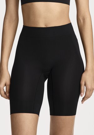 Woman wearing high-waisted black bike shorts and a black crop top with hands relaxed at sides against a white background.