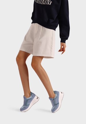 Sneakers blu con una combinazione di tessuto liscio e strutturato, dotate di zip e suola bianca imbottita, abbinate a pantaloncini chiari.