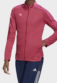 Chaqueta deportiva rosa con cierre frontal, cuello alto y acentos de tres rayas en las mangas; textura suave con bolsillos laterales y detalle de marca.