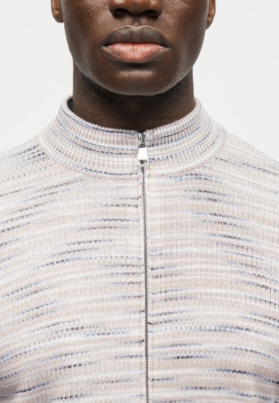 Gros plan d'une personne portant un pull zippé côtelé de couleur claire avec des lignes horizontales subtiles bleu et gris.