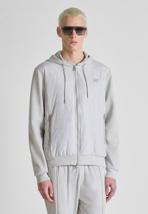 Jonge man met kort blond haar, grote donkere zonnebril, lichtgrijze hoodie met rits en bijpassende broek, staand tegen een witte achtergrond.