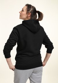 Felpa nera con una vestibilità comoda, dotata di cappuccio, maniche lunghe e polsini a costine. Indossata con pantaloni grigi, presenta un design semplice.