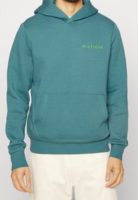 Tyrkysová mikina s kapucí, která má přední kapsu, žebrované manžety a zelené logo "HILFIGER" na hrudi. Měkký, hladký bavlněný materiál.