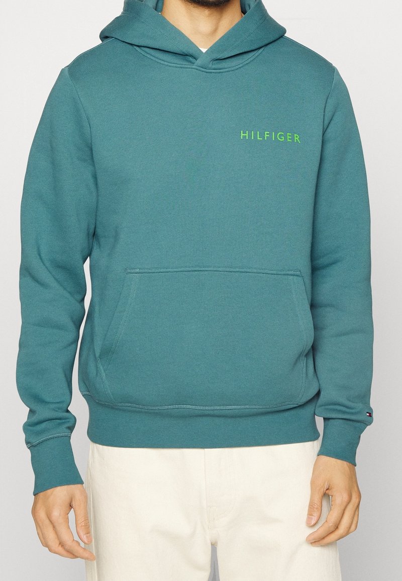 Tyrkysová mikina s kapucí, která má přední kapsu, žebrované manžety a zelené logo "HILFIGER" na hrudi. Měkký, hladký bavlněný materiál.
