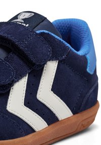 Marinblå mockaskor med vita zigzagdetaljer, kardborreband och gummisula. Inuti finns en blå meshfoder.