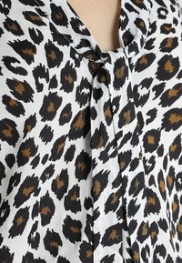 Blus med leopardmönster i vitt tyg, med svarta och bruna fläckar, en krage med en knut vid halsen och en slät textur.