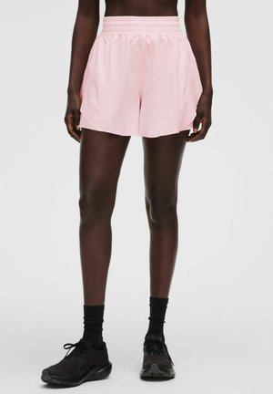 Person iført lyserøde sportsshorts, sorte sokker og sorte sneakers, stående mod en ensfarvet lys baggrund.