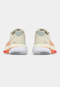 Beige Sportschuhe mit einer strukturierten Oberseite und orangefarbenen Streifen. Feste weiße Sohle mit einem Farbverlauf an der Ferse, akzentuiert durch eine hellgraue Fersenlasche.