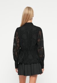Černý peplum top s květinovou průhlednou tkaninou, dlouhými rukávy, elastickým pasem a knoflíkovými manžetami, spárovaný s skládací sukní tmavě šedé barvy.
