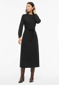 Černé pletené šaty s dlouhými rukávy, kulatým výstřihem a látkovým pásem. Sukně je dlouhá až ke kotníkům, doplněná lesklými hnědými kozačkami ke kolenům.