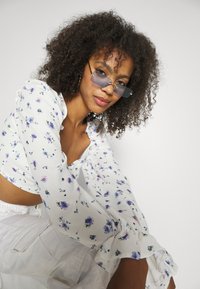 Blusa floral branca com realces roxos, mangas longas e punhos com babados; combinada com óculos de sol metálicos e coloridos.