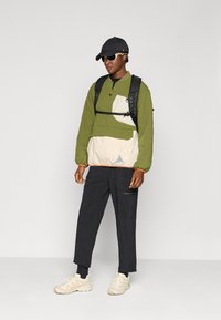 Chaqueta de pullover con bloques de color verde y crema, con cremallera, pantalones negros, zapatillas crema y una gorra negra. Equipado con una mochila negra.