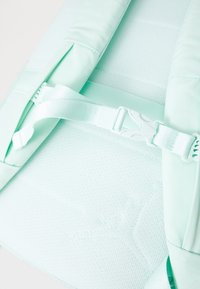 Gaišas mentolzaļas mugursomas detaļas ar sietu audumu, mīkstinātām siksnām un plastmasas regulēšanas sprādzēm. Gluda tekstūra un minimālisma dizains.