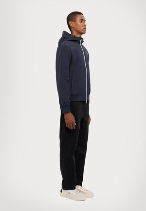 Giacca a vento blu marina con cappuccio e zip bianca, dotata di polsini elasticizzati, abbinata a jeans neri e sneaker bianche.