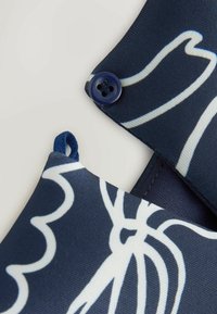 Tissu bleu marine avec motifs floraux abstraits blancs, présentant une petite boucle et un bouton à quatre trous cousu sur les coins superposés.