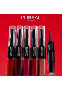 Six rouges à lèvres L'Oréal Paris Infallible 24HR dans des tubes transparents, présentant des teintes de rouge, rose et nude, avec un applicateur noir à droite.