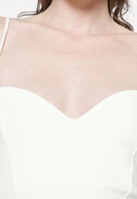 Femme portant une robe blanche sans bretelles montrant le haut de sa poitrine et ses clavicules sur un fond uni.