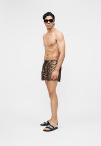 Hemdloser Mann trägt Schwimmshorts mit Leopardenmuster, schwarze Sonnenbrille und schwarze Slides mit weißem "Moschino"-Schriftzug vor weißem Hintergrund.