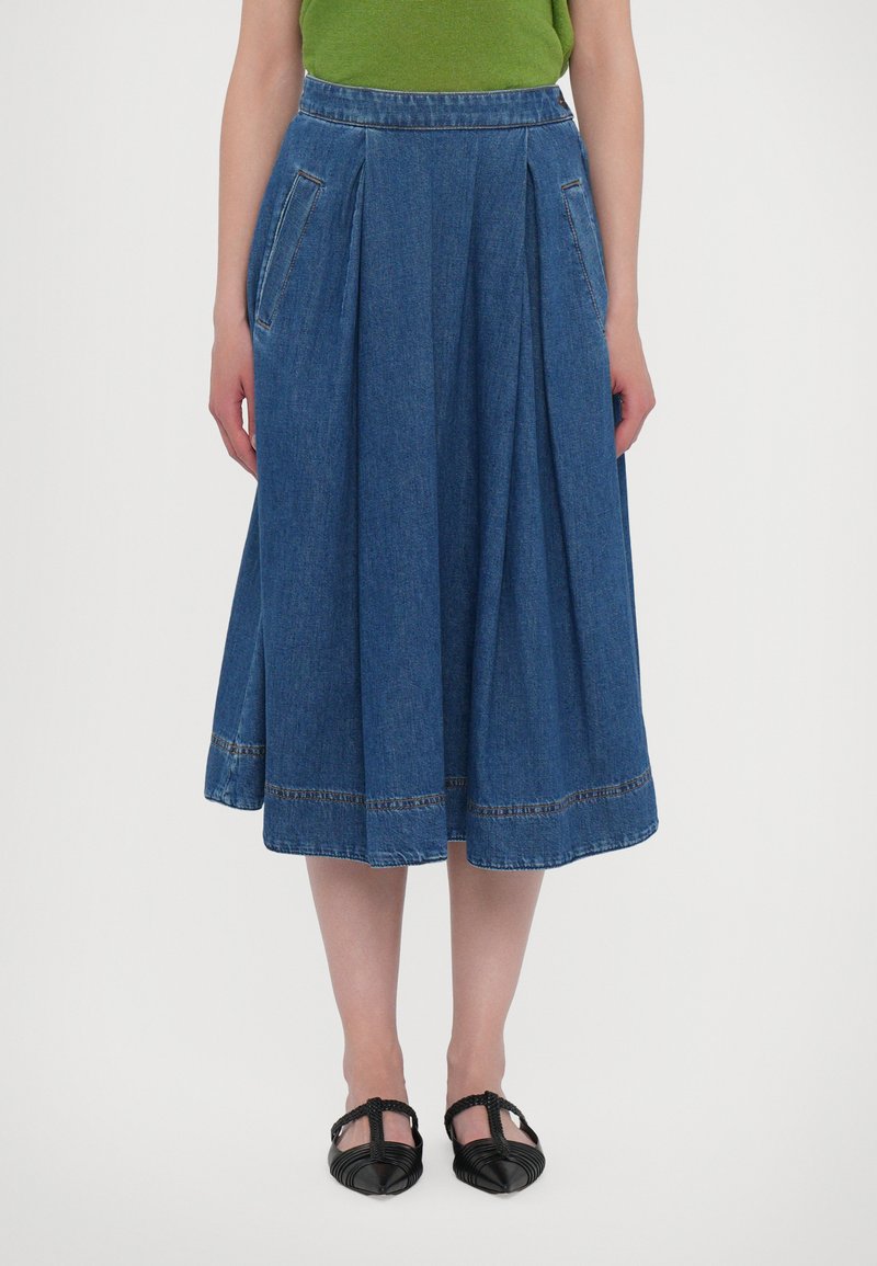 Denim rok met een uitlopende silhouet, voorzien van twee zijzakken, plooien en een zoom met contrasterende stiksels. Draagt met zwarte platte schoenen.