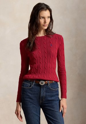 Femme portant un pull en maille torsadée rouge, un jean bleu et des bottines beiges, debout devant un fond neutre.
