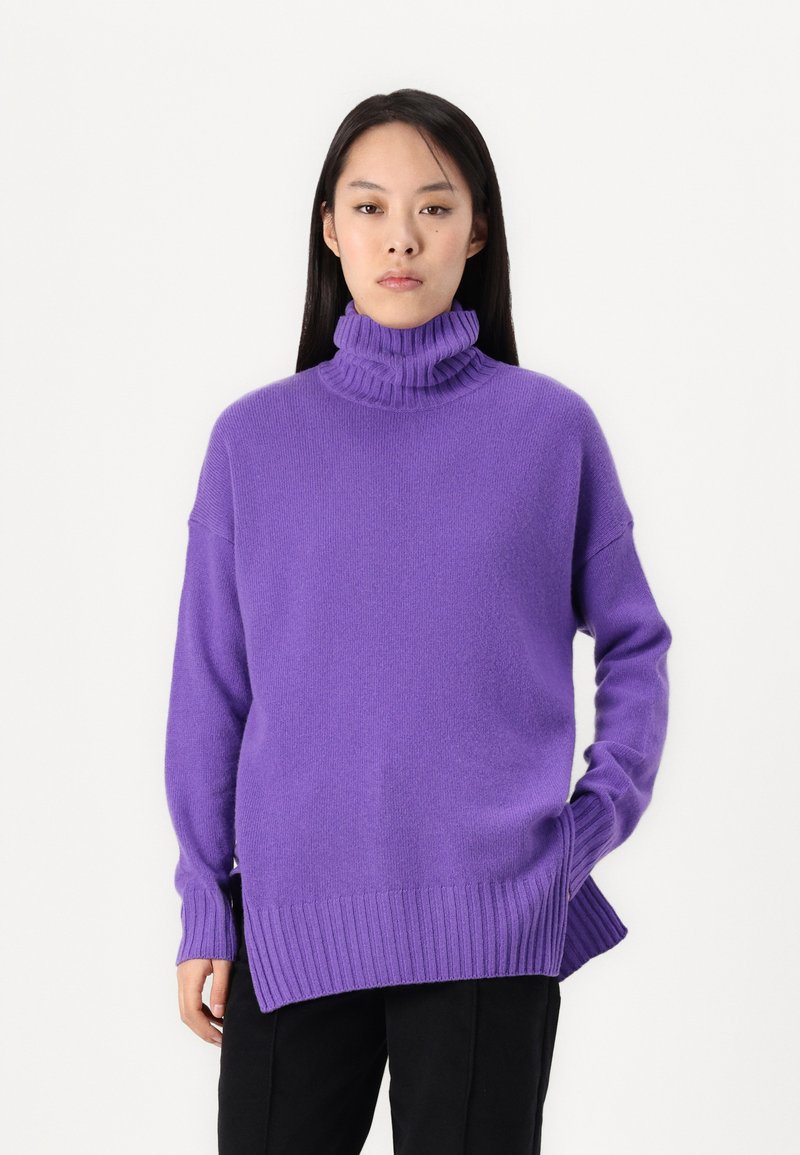 Lila, gerippter Rollkragenpullover mit lockerem Schnitt, seitlichen Schlitzen und langen Ärmeln, aus einem weichen Material. Kombiniert mit schwarzen Hosen.