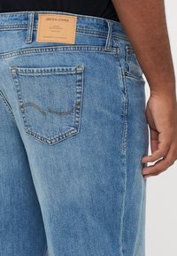 Zbliżenie osoby w jasnoniebieskich dżinsach Jack & Jones, pokazujące tylną kieszeń i metkę marki na pasku.