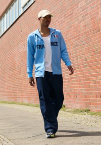 Hellblaue Zip-Jacke mit dem Aufdruck "FRANCE", weißes T-Shirt, dunkelblaue weite Jeans und schwarze Sneakers mit grünen Akzenten.