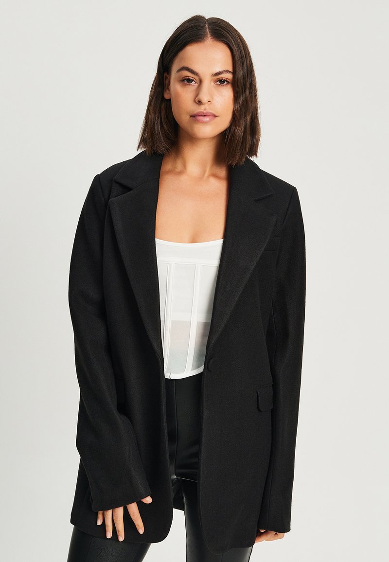 Blazer noir ajusté avec un col à revers, fermeture à un bouton et deux poches avant. Tissu texturé, coupe décontractée et manches longues.