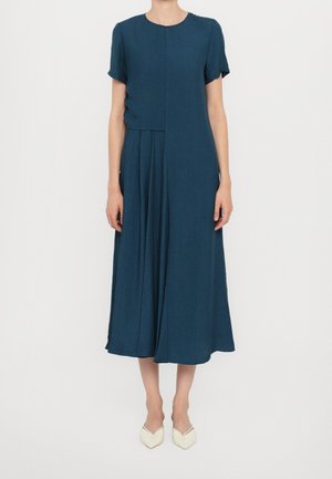 Woman standing wearing a dark teal short-sleeve midi dress with pleats and white pointed flats on a plain white background.